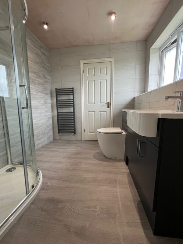 Light grey tiled bathroom, dark grey long vanity unit that houses both the basin and the toilet, dark grey ladder towel warmer, rounded corner shower enclosure.